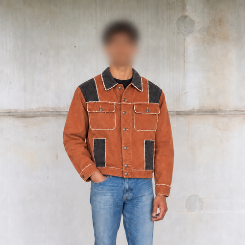 A person wearing a rust-orange denim jacket with black contrast panels and light piping, paired with blue jeans and black shoes, standing against a concrete wall.