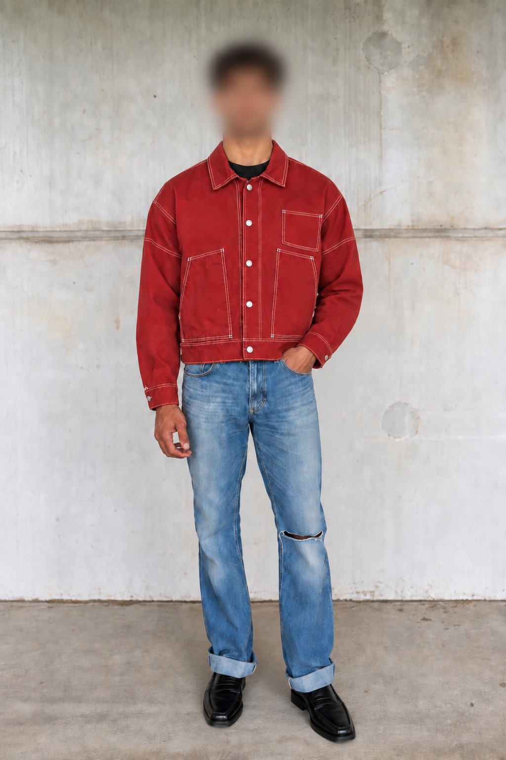 A person wearing a red denim jacket over a black shirt, blue jeans, and black shoes, standing against a textured concrete wall.