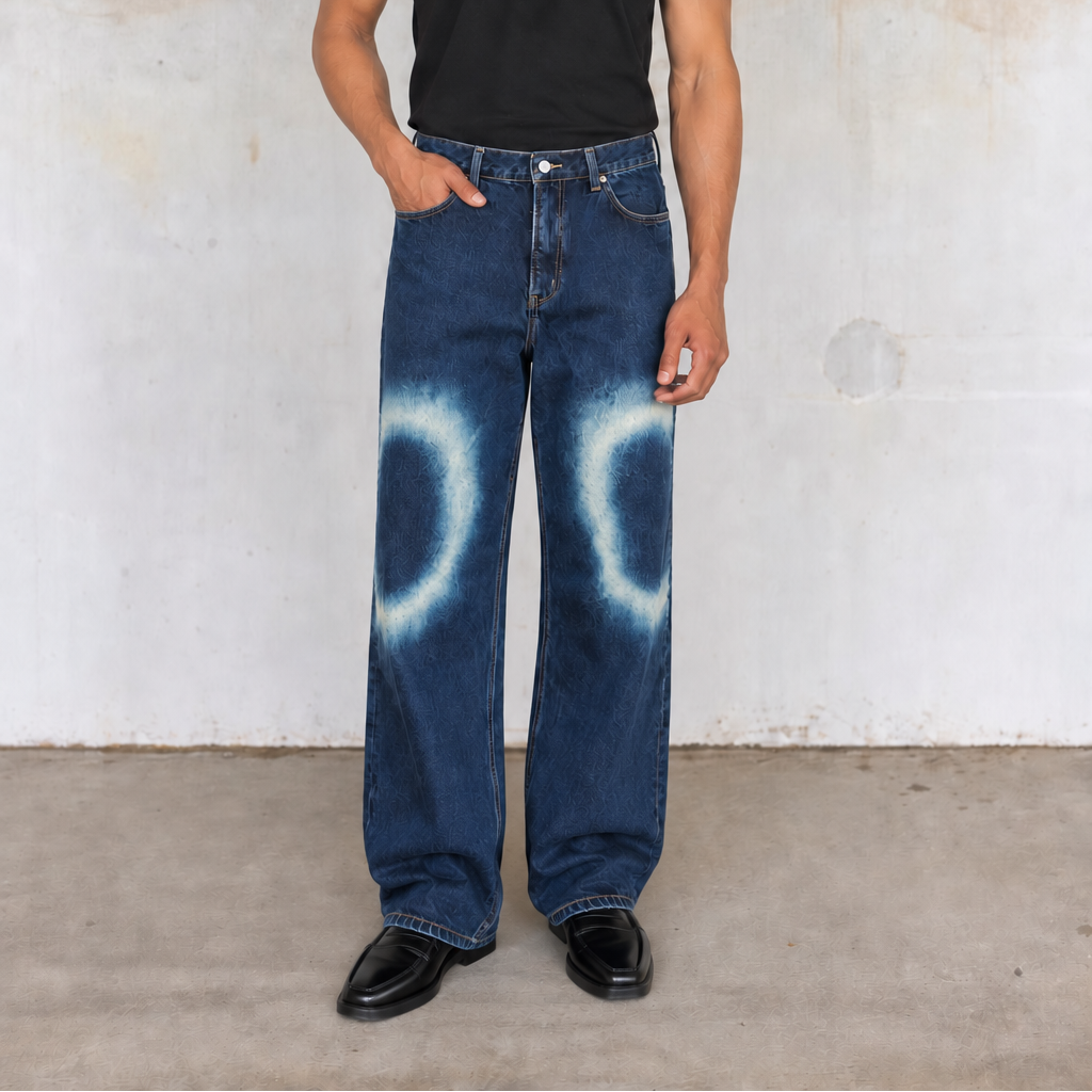 A person stands against a textured concrete wall wearing loose dark blue jeans with a white tie-dye circular pattern on the lower legs, a black short-sleeve t-shirt, and black shoes.