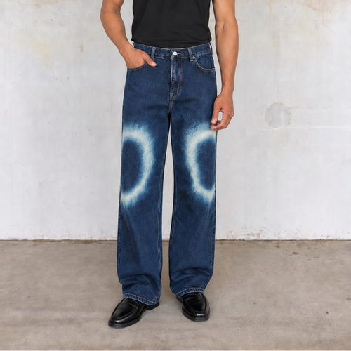 A person stands against a textured concrete wall wearing loose dark blue jeans with a white tie-dye circular pattern on the lower legs, a black short-sleeve t-shirt, and black shoes.