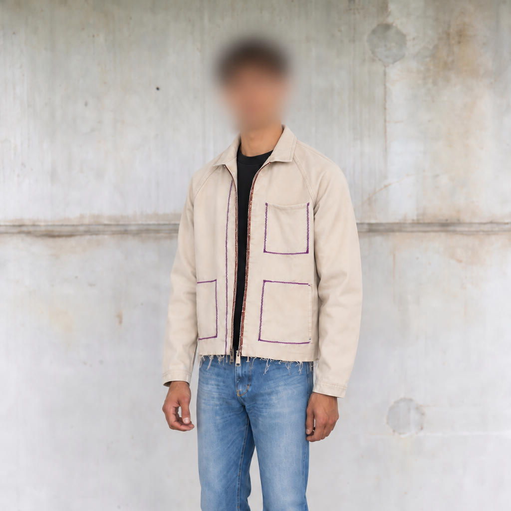 A person stands against a textured concrete wall wearing a light beige jacket with purple piping details, blue jeans, a black shirt, and black shoes.